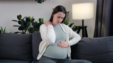 Unwell young Caucasian pregnant woman suffer from neck pain ache sitting on sofa at home. Unhealthy tired brunette female have health problems during pregnancy, struggle with backache or muscle spasm
