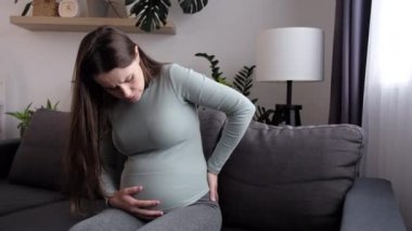 Upset young pregnant woman massaging back muscles sitting on couch at home, suffering from severe painful feelings, future mom with third trimester big belly having joints strain in lower back