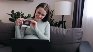 Happy beautiful young pregnant woman showing photo of baby during online call with family by pc laptop while sitting on cozy sofa. Smiling future mother talking via video link from pc computer at home