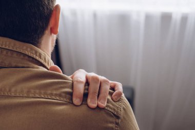Side view of unhealthy young man sitting near window suffering from pain in shoulder, doing self-massage. Neck pain. Forearm pain, people with body muscle problems, healthcare and medicine concept