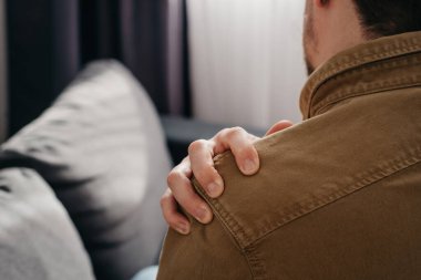 Pain in shoulder. Upper arm pain, people with body-muscles problem, healthcare and medicine concept. Close up back view of young man sitting on sofa and holding painful shoulder with another hand