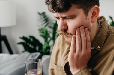 Sad caucasian male 30s sit on couch at home touching cheek, feel hurt and suffering from sensitive tooth ache. Pain and cavities concept. Upset young man feel terrible toothache after drink cold water