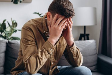 Close up of depressed young bearded man sitting on grey couch in living room, leaning on hands, having financial troubles during quarantine or suffering from loneliness. Depression and sadness concept