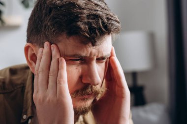 Closeup of upset young caucasian man suffering from headache sitting on couch at home, touching his temples. Migraine, headache, mental stress, panic attack, tension problem, hangover concept