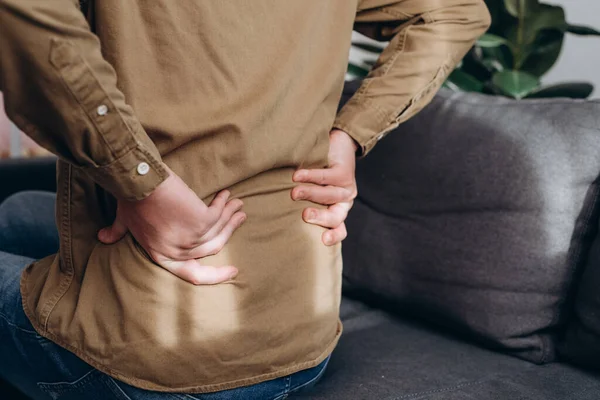 Side view of unhealthy young caucasian man sitting on couch at home suffering from back pain, have backache, health problems, lower back, has chronic joint problems. Problems with spine concept