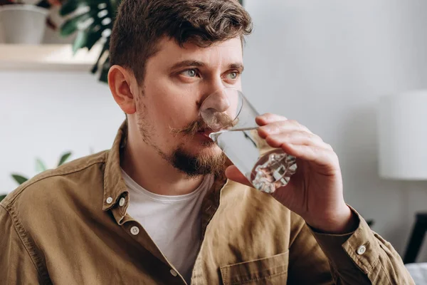 Close up of young bearded man drinking fresh water from glass, sitting on sofa in living room at home. Health care, proper nutrition at home, thirsty in morning and domestic life, hydration concept