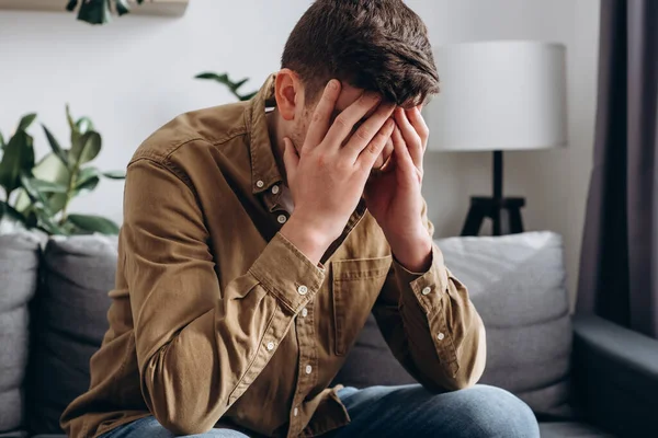 Close up of depressed young bearded man sitting on grey couch in living room, leaning on hands, having financial troubles during quarantine or suffering from loneliness. Depression and sadness concept