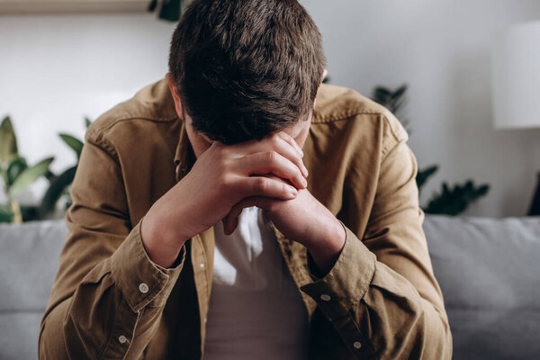 Depression and sadness concept. Close up of depressed young caucasian man sitting on sofa in living room, leaning on hands, having financial troubles during quarantine or suffering from loneliness