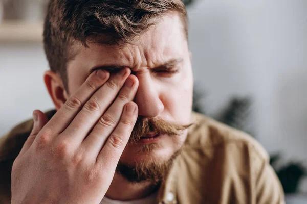 Selective focus of frustrated sad caucasian man suffering from strong eye pain. Eyesight problem. Upset young bearded male 30s touching eyes feeling discomfort. Injuries poor health Illness concept