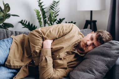 Sad young man lying on grey couch at home, suddenly feeling strong stomach ache, gastritis problem. Upset male squeezing belly with hands because of abdominal pain. Guy suffering from stomach ache