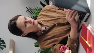 İş yerindeki yorgun genç kadının dizüstü bilgisayarda çalıştığı ve parmaklarında ağrı hissettiği dikey bir video, kız esnemeye çalışır ve eklemlerine tıklamaya başlar. Acı ve parmak şıklatma. Eklem iltihabı