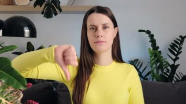 Oturma odasındaki kanepede oturan esmer kadın başparmağının yakın portresi. Ciddi bir beyaz kadın kameraya bakıp el kaldırıyor ve baş parmağını aşağı gösteriyor. İnsanların duygu kavramı