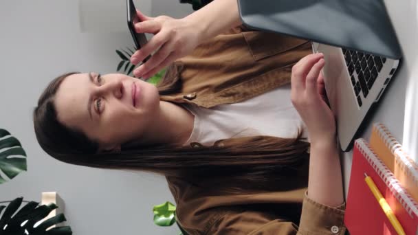 幸せな若い女性の垂直ビデオは 自宅のオフィスでコンピュータ上でオンラインで動作し スマートフォン上で音声メッセージを記録します かわいい笑顔の女性従業員は 携帯電話上のスピーカーでノートパソコンの話で購入 — ストック動画