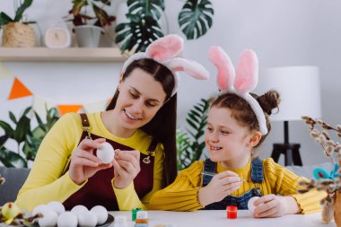Smiling young mother teaching pretty little daughter child painting eggs sitting together at table, wears bunny ears. Easter celebration and traditions. Concept of creativity and children's education