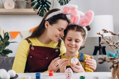 Loving young mother hugging pretty little daughter child, happy family painting eggs sitting at table, wears bunny ears. Easter celebration and traditions. Concept of creativity and spring holiday