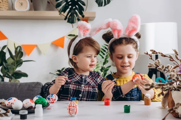 Spring holidays and family concept. Cheerful cute little sisters in pink fluffy bunny ears preparing for Easter together, children painting eggs with paints and brushes while sitting at white table
