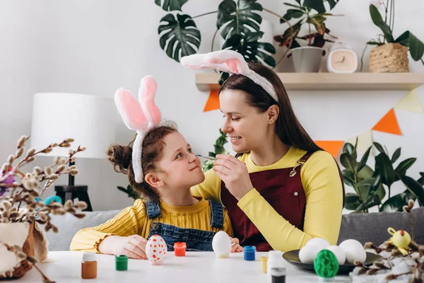 Easter Family traditions concept. Positive young mother and smiling little daughter to dye and decorate eggs with paints for Easter holidays, play, laugh and draw on faces while sit together at table