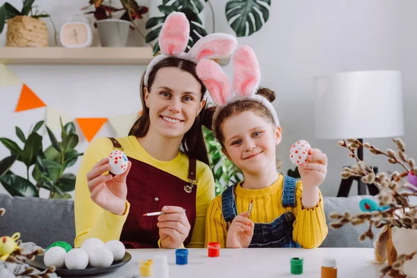 Sweet family portrait of smiling young mom and pretty little daughter in fluffy bunny ears holding small painted multi-colored Easter eggs, tenderly embracing and smiling in cozy living room at home