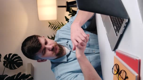 疲れた青年の垂直ビデオはノートパソコンでテーブルに座って指の痛みを経験し 男性は引き伸ばそうとし 関節をクリックし始めます 痛みや指をスナップ 関節炎の概念 — ストック動画