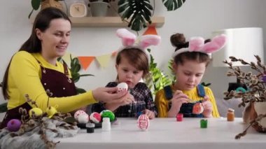 Mutlu bir anne ve iki küçük kız Paskalya tatili için yumurtaları boyayarak dekore masasında birlikte oturuyorlar. Tavşan kulağı takan komik bir aile Paskalya günü yumurta boyuyor.