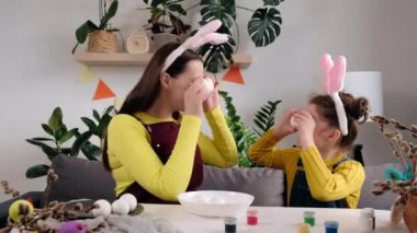 Mutlu, genç bir anne ve neşeli küçük bir kız çocuğu beyaz Paskalya yumurtalarını gözünün önünde tutarken onları evdeki rahat odada yemek boyalarıyla süslüyorlar. Aileler için Paskalya Sanatları etkinlikleri