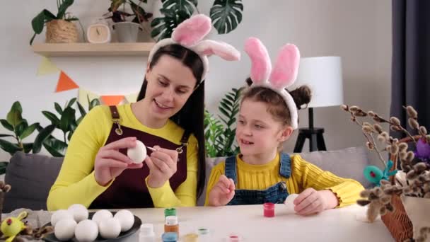 イースターのお祝いと伝統 テーブルに座っている卵を描き ウサギの耳を身に着けているかわいい娘の子供の絵を教えて笑顔の若い母親 創造性と子供の教育の概念 — ストック動画