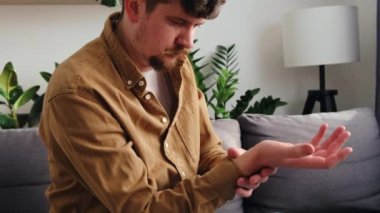 Ellerinde ve parmaklarında ağrı, eklem iltihabı olan üzgün, genç, beyaz bir adama yakın durun. Üzgün sakallı erkek, evdeki kanepede otururken bileğinde ağrı hissediyor. Sağlık ve tıbbi konsept