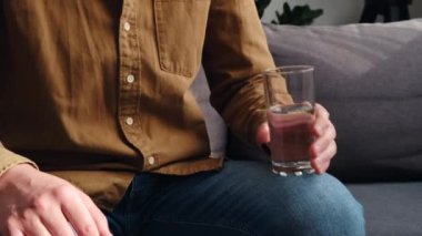 Genç sakallı adamın bardaktan tatlı su içtiği seçici odak noktası, evdeki oturma odasında kanepede oturmak. Sağlık hizmeti, evde düzgün beslenme, sabah ve ev yaşamında susuzluk, hidrasyon konsepti.