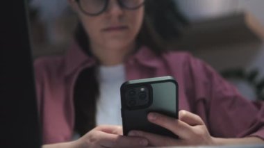 Genç bayanların seçici odak noktası çalışmak veya çalışmak için cep telefonu uygulaması ve bilgisayar kullanmaları, karanlık odada oturmaları. Gözlüklü bir kadın cep telefonu ekranına bakıyor ve ofiste dizüstü bilgisayar kullanıyor.