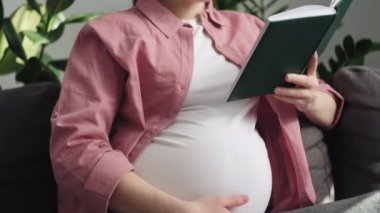 Gelişmiş hamilelik dönemindeki mutlu genç bir kadın rahat koltuğunda oturup kitap okuyarak, karnını nazikçe okşayarak, bebek tekmelerini hissederek rahatlıyor. Annelik kavramı