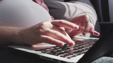 Müstakbel annenin bilgisayarlı hamile karnının yakın çekimi. Arama, çevrimiçi iletişim, internet üzerinden alışveriş ya da hamilelik ve doğum izni sırasında çalışma. Hamile kadın evde dizüstü bilgisayar kullanıyor.