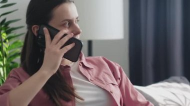Gülümseyen çekici, hamile bir kadın akıllı telefondan konuşuyor. Evdeki kanepede oturuyor. Müstakbel annenin oturma odasında cep telefonuyla koltukta oturması. Müstakbel anne göbeğini okşuyor