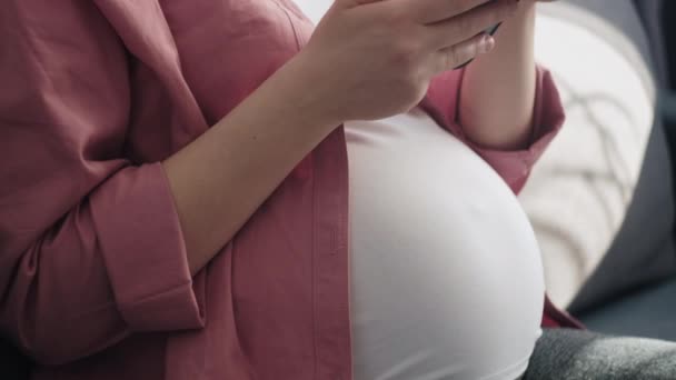 快乐美丽的年轻孕妇坐在舒适的沙发上 坐在明亮的客厅里 使用智能手机 手指触摸屏幕 宝宝的预产期家庭放松和怀孕的概念 — 图库视频影像