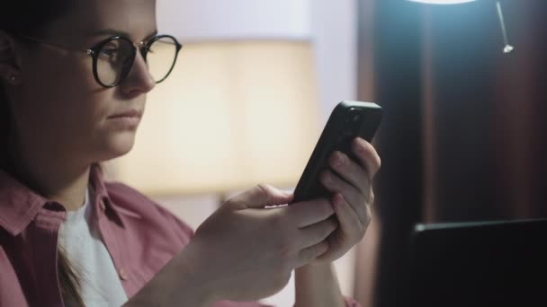 眼鏡の若い女性の選択的な焦点は 携帯電話を使用してソーシャルメディアでテキストメッセージを共有するテーブルに座って ノートパソコン上での作業からインターネットコンテンツを見てください 電子サービス モバイルアプリの使用の概念 — ストック動画