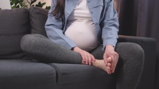 妊娠後期の白人女性の自宅で灰色のソファに座って閉じます 痛みを伴う足首をマッサージ 期待される母親の太りすぎの疲れ 健康上の問題を持っています 関節痛の概念 — ストック動画