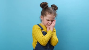 Üzgün küçük kız yanağa dokunuyor, acı dolu diş ağrısı, hassas dişler, çürükler yüzünden gözleri kapatıyor stüdyonun mavi arka plan duvarına izole edilmiş poz veriyor. Diş problemi