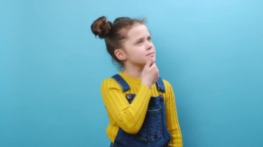 7-8 yaşlarında, sıradan kıyafetleri içinde, parlak bir fikirle işaret parmağıyla stüdyodaki mavi arka plan duvarına izole edilmiş bir şekilde poz veren düşünceli küçük bir kız çocuğu. Çocukluk yaşam tarzı, zekice bir kavram.
