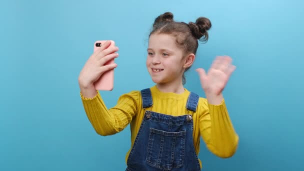 快乐的小女孩博主拿着智能手机自拍 与订户在网上进行视频通话 在工作室的蓝色背景墙上 漂亮的孩子孤身一人摆姿势 — 图库视频影像
