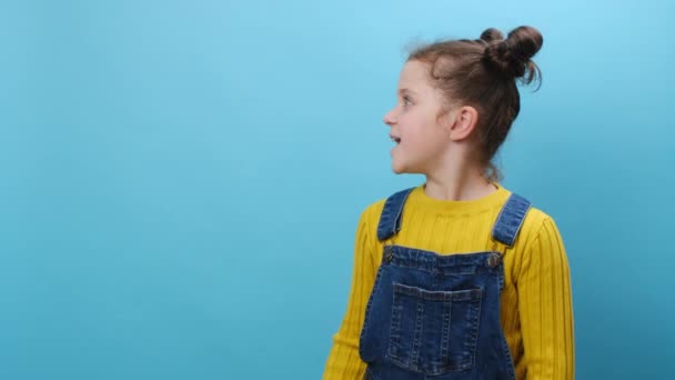 兴奋的小女孩把食指放在一边 像个手势一样伸出大拇指 在工作室蓝色的背景墙上摆出孤立无援的姿势 童年生活方式和人的情感概念 — 图库视频影像