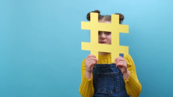 在工作室里 一个可爱的 嬉闹的小女孩拿着黄色的Hashtag标志 开心地看着镜头 脸上挂着哈希牌 在蓝色的背景墙上摆出一副与世隔绝的样子 — 图库视频影像