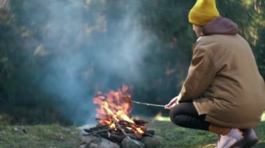 Bahar ya da yaz günü kamp ateşinde ekmek yapan genç kadın turistlerin seçici odak noktası. Dişi gezgin yeşil ormanda şenlik ateşinde yemek pişiriyor. Kamp ortamı ve açık hava yaşam tarzı konsepti