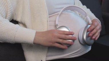 Hamile bir kadının karnına kulaklık takıp evdeki rahat koltukta yalnız başına oturmasına yakın çekim. Bekleyen anne oturma odasında müzik dinliyor. Müstakbel anne doğmamış çocuklara müzik veriyor.