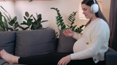 Kablosuz kulaklıkla müzik dinleyen rahat, mutlu, hamile bir kadın. Evdeki oturma odasında rahat bir kanepede dinlenirken akıllı telefon kullanan kaygısız müstakbel bir anne. Hamilelik, hobi konsepti