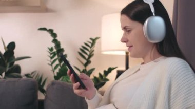Cep telefonuyla bağlı kulaklık takan, akıllı telefondan müzik dinleyen güzel bir anne. Mutlu, güzel, hamile, genç bir kadın evde boş vakit geçiriyor. Melodi terapi konsepti