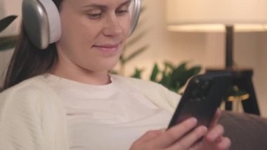 Evdeki rahat koltukta oturmuş kulaklıkla müzik dinleyen mutlu, tatlı hamile kadının seçici odak noktası hamilelik süresince rahatlama ve kişisel bakım, akıllı telefon ve teknoloji, hamilelik ve rahatlama.