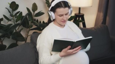 Kulaklık takan, müzik dinleyen, evde rahat bir koltukta oturup kitap okuyan çekici hamile bir kadın. Güzel müstakbel anne teknolojinin tadını çıkarıyor. Hamilelik konsepti