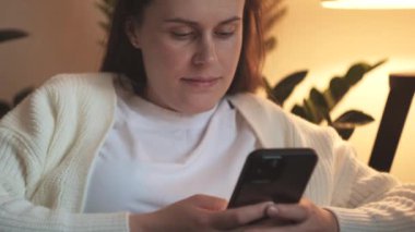 Oturma odasındaki rahat koltukta oturan, cep telefonu kullanan, boş zamanlarını okuyarak ve sosyal ağları kontrol ederek geçiren çekici, sakin, genç, beyaz bir kadın. Teknoloji konsepti