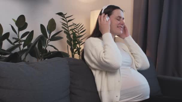 幸せな美しい期待の母親音楽を聴いてメロディー楽しい妊娠座っている居心地の良いソファに自宅で 取り上げヘッドフォンから頭を置く妊娠中の腹のオーディオ療法 Unborn Baby — ストック動画