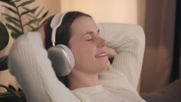 积极的年轻孕妇在无线耳机听音乐 虚拟播放清单的压力缓解和放松 享受闲暇的孕妇坐在舒适的沙发上 怀孕的概念 — 图库视频影像