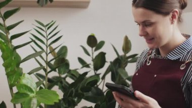 Gülümseyen genç bir kadın kanepede otururken önlük giyer akıllı telefon kullanır ve evdeki kapalı salonda çevrimiçi dükkanda satmak için bitki yapraklarını kontrol eder. Yaşam tarzı, bahçıvanlık ve aktivite konsepti